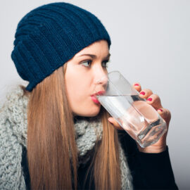 mulher com roupa de frio, toca e cachecol tomando um copo de água